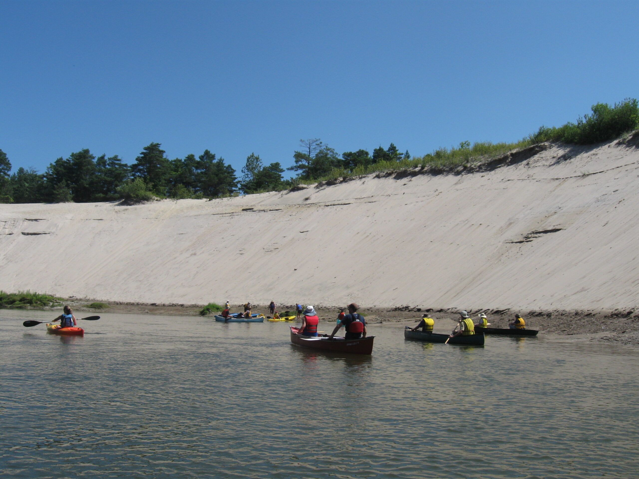 Canoes paddling in a river beside a large hill of sand